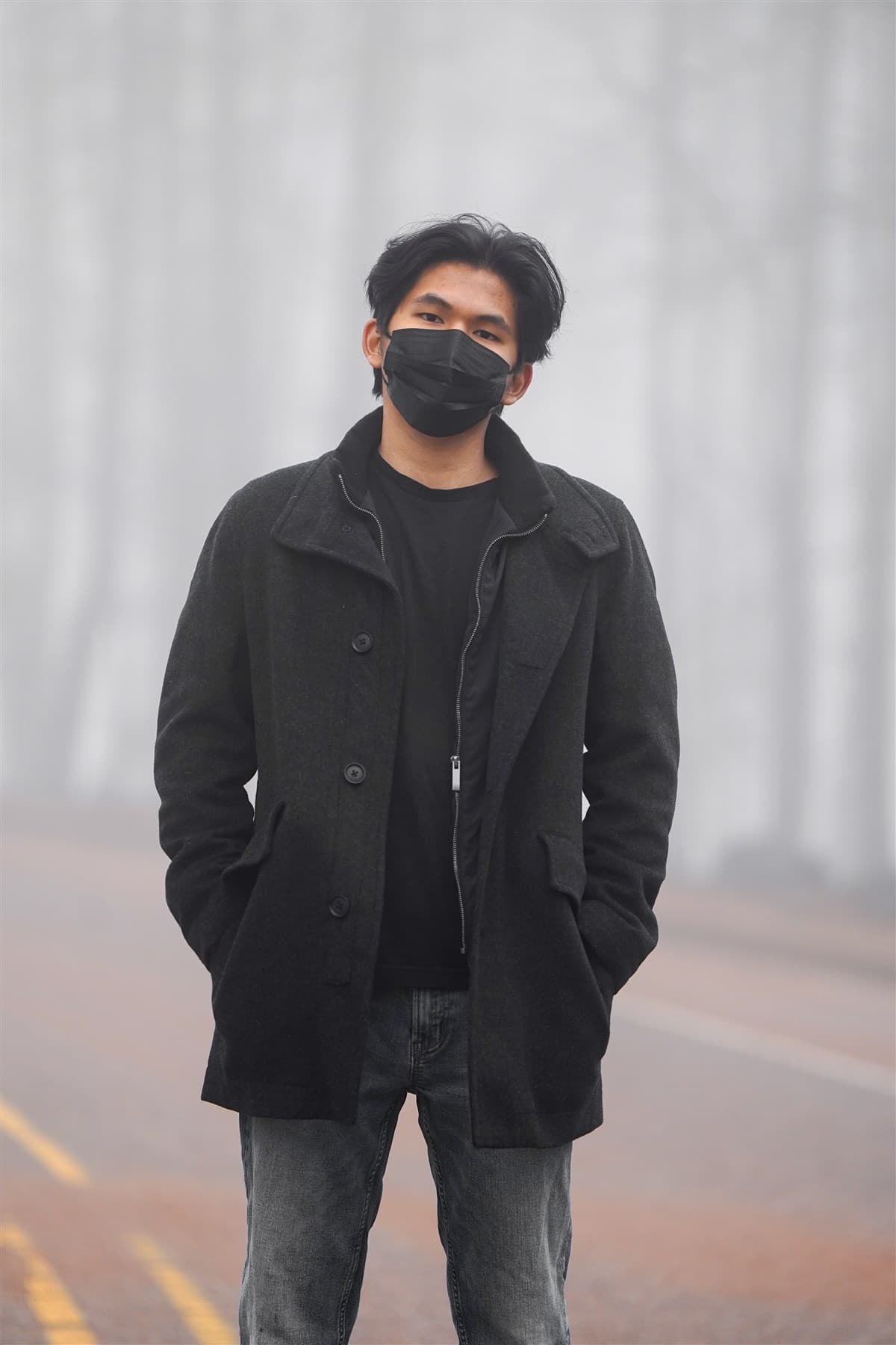 Portrait of the photographer standing on a foggy road in a black coat and mask.